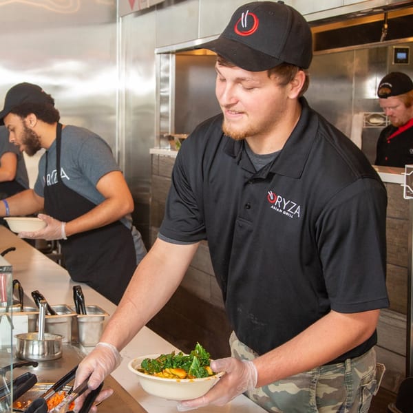 Oryza Asian Grill employee in a black polo shirt and hat preparing a fresh bowl of food.