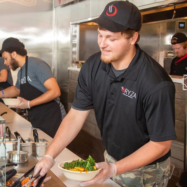 An Oryza Asian Grill employee in a black polo shirt and hat prepares a fresh bowl of food.