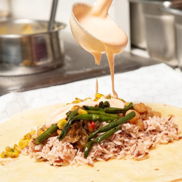 A ladle pours creamy sauce over a burrito filled with rice, green beans, corn, and shrimp.