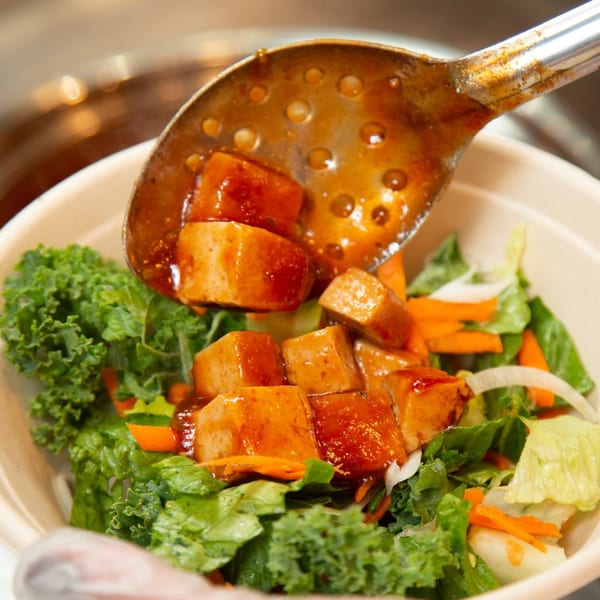 A slotted spoon adds glazed tofu to a fresh salad with kale, lettuce, carrots, and onions.