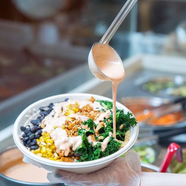 A ladle pours creamy sauce over an Oryza Asian Grill bowl with kale, corn, black beans, and crispy toppings.