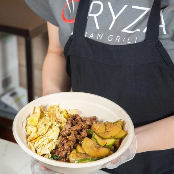 A person in an Oryza Asian Grill apron holds a bowl with scrambled eggs, beef, and zucchini.