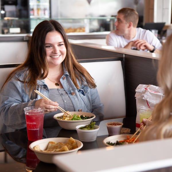 A smiling woman in a denim jacket eats an Asian-fusion bowl with chopsticks at Oryza Asian Grill.